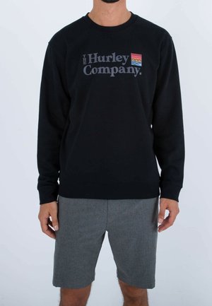 Uomo che indossa una felpa nera con il logo "The Hurley Company" e un piccolo emblema colorato, abbinata a pantaloni corti grigi, in piedi di fronte a uno sfondo bianco.