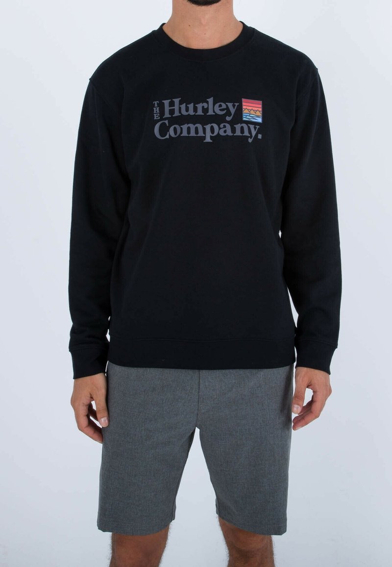 Muž v černé mikině s logem "The Hurley Company" a malým barevným emblémem, oblečený v šedých šortkách, stojí na bílém pozadí.