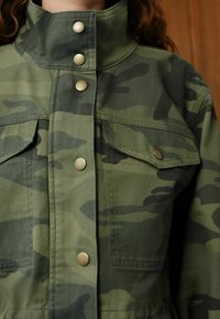 Gröntonad jakke med camouflagemönster, har hög krage, knappknäppning och två framfickor med lock. Tillverkad av hållbart tyg.
