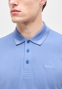 Άνδρας μοντέλο που φοράει ανοιχτό μπλε πόλο με κουμπιά και γιακά, με το λογότυπο "BOSS" κεντημένο στο στήθος.