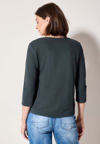 Haut texturé vert foncé à manches trois-quarts, col rond et détails de boutons sur les manches, associé à un jean bleu clair.