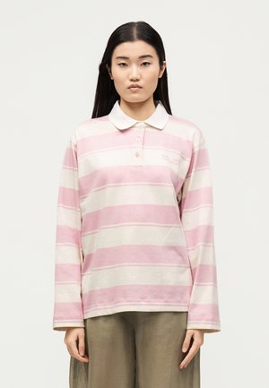 Ung kvinde med langt sort hår iført lyserød og hvidstribet langærmet polo og løse lysebrune bukser, stående mod hvid baggrund.