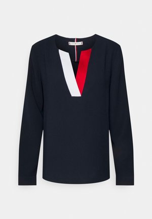 Marineblå bluse med lange ærmer og V-udskæring med røde og hvide farveblokke på venstre og højre side af udskæringen.