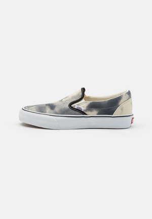 Slip-on sneakers med en spräcklig grå och krämfärgad canvasoversida, svarta elastiska sidodetaljer, vit gummisula och Vans-logotyp.