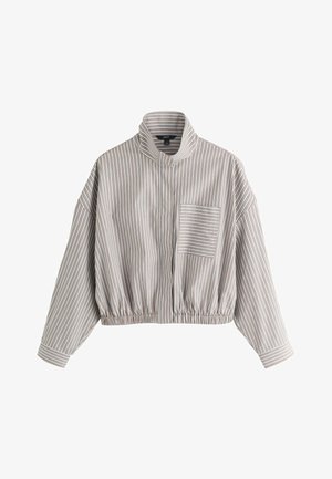 Jeune femme aux longs cheveux blonds et lunettes de soleil noires, portant une chemise rayée, se tient devant un paysage urbain avec des toits en terre cuite.