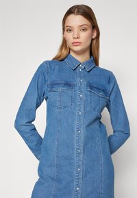 Rochie din denim de culoare albastră deschisă. Dispune de o guler ascuțit, două buzunare pe piept, închidere cu nasturi și mâneci lungi. Textură netedă.