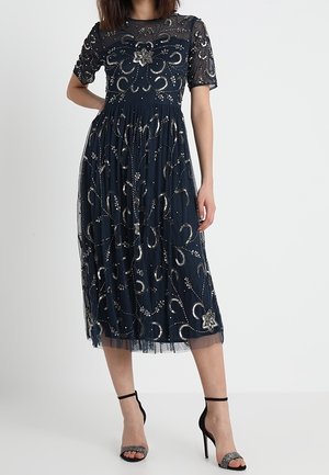 Granatowa sukienka midi z krótkimi rękawami, przezroczystą nakładką, cekinowymi zdobieniami, kwiatowymi detalami i plisowaną spódnicą. Do kompletu sandały na cienkich paskach.