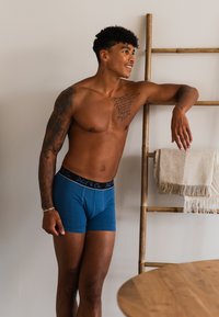 Modré bavlněné boxerky s černým elastickým pasem, se značkovým logem. Model se opírá o dřevěný žebřík a ukazuje tetování.