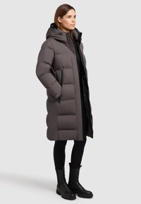 Grey puffy coat with a hood, featuring a smooth, quilted texture, zippered pockets, and a long silhouette. Worn over black leggings and boots.