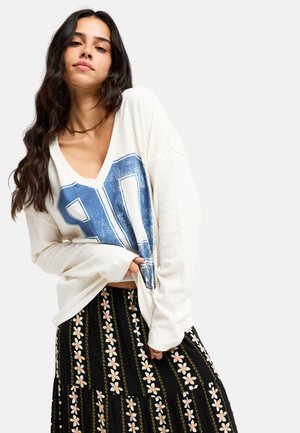 Femme aux longs cheveux foncés portant un sweat-shirt blanc oversize avec un imprimé chiffre bleu et une jupe noire à motif floral.