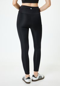 Zwarte legging met een hoge tailleband, een gladde textuur en zijnaadjes. Gecombineerd met witte sneakers met groene strepen en een bruine zool.