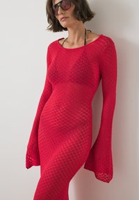 Punane kootud kleit, millel on pikad, laienevad varrukad ja avatud teemantkude. Kerge ja keha ligi, näidates materjali all olevat nahka.