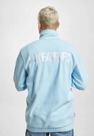 Lühikeste blondide juustega inimene, kes kannab seljal tekstiga „UNFAIR FC“ hele sinist fliisjopet, seisab tavalise tausta ees.