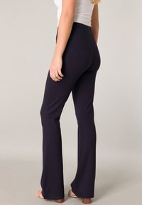 Marineblaue Schlaghosen aus einem dehnbaren Material mit hoher Taille, glatter Textur und sauberen Nähten, kombiniert mit hellfarbigen Sandalen mit Blockabsatz.