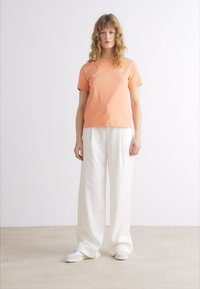 Maglietta arancione a maniche corte con scollo tondo, abbinata a pantaloni bianchi larghi e sneaker bianchi su uno sfondo neutro.