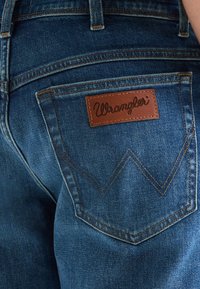 Tylna kieszeń niebieskich dżinsów z brązową skórzaną naszywką marki Wrangler i podwójnym przeszyciem w kształcie litery "W".