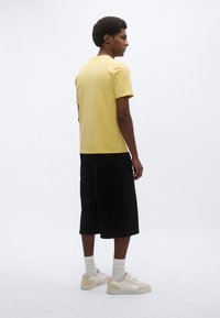 Κίτρινο κοντομάνικο μπλουζάκι, μαύρα σορτς μέχρι το γόνατο, λευκά αθλητικά παπούτσια και λευκές κάλτσες. Απλός σχεδιασμός με χαλαρή εφαρμογή, χωρίς εμφανή σχέδια.