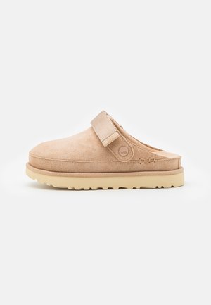 Sabot beige en daim à enfiler avec sangle réglable et semelle en caoutchouc texturée, conçu pour un usage décontracté en intérieur comme en extérieur.
