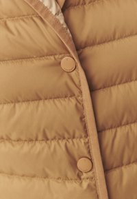 Doudoune beige avec sections matelassées horizontales, présentant une texture lisse et une petite fermeture à bouton rond. Design minimaliste.