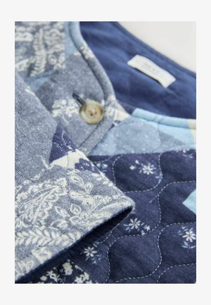 Tikkidega kangas, millel on siniste toonide segu ja valged lillemustrid. Üksikasjadesse kuuluvad nupp ja kontrastne krae.