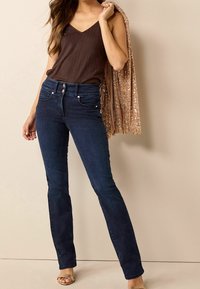 Jean en denim foncé à taille haute avec un design à cinq poches, associé à un débardeur marron côtelé et un châle à paillettes.