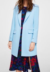Donna che indossa un cappotto su misura blu chiaro sopra un vestito floreale scuro con fiori rossi, blu e verdi, in piedi davanti a uno sfondo semplice.