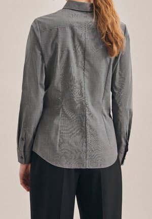 Donna con capelli lunghi che indossa un blazer grigio aderente e pantaloni neri, vista da dietro su uno sfondo neutro.
