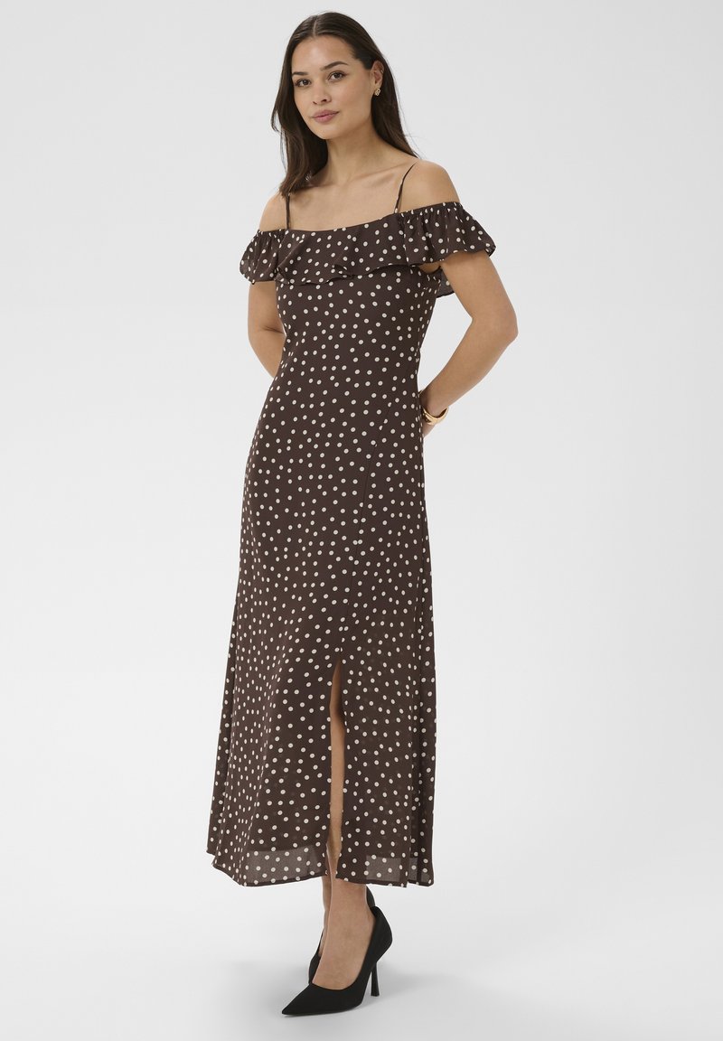 Femme debout, mains derrière le dos, portant une robe marron à pois décolletée avec fente à l'avant et des talons hauts noirs sur fond blanc.