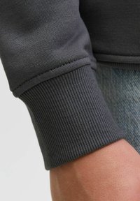 Poignet de sweat-shirt gris présentant une texture côtelée et des bords cousus, associé à un fond en denim clair.