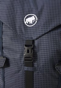 Blå mønstrete ryggsekk laget av slitesterkt stoff. Har en hvit logo, strukturert overflate og en svart spenne med justerbart webbing.