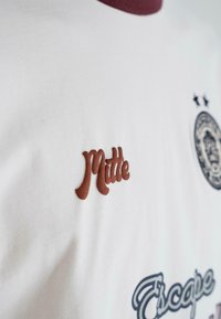 Balts krekls ar brūnu tekstu "Mitte", apaļu emblēmu ar divām zvaigznēm un stilizētu tekstu "Escape" zilā un pelēkā krāsā.