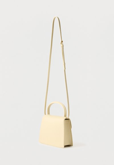 Μικρή μπεζ δερμάτινη τσάντα με χειρολαβή στο πάνω μέρος και μακρύ ρυθμιζόμενο λουρί ώμου, σε απλό φωτεινό φόντο.