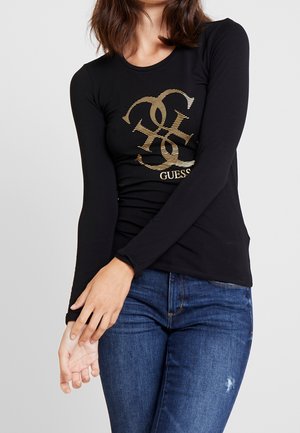 Femme portant un t-shirt noir à manches longues avec le logo doré Guess et un jean bleu, posant avec les bras croisés devant un fond gris clair.
