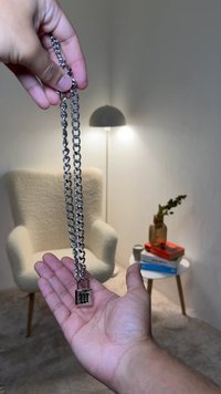 Corrente de metal prateada com um pendente de cadeado, apresentando um design entrelaçado. Uma mão segura a corrente acima de uma cadeira e uma mesa de cor clara.