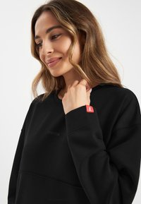 Czarny sweatshirt z teksturowanym wzorem, subtelnie wytłoczonym logo oraz akcentem w postaci czerwonej metki na rękawie. Miękka tkanina, luźny krój.