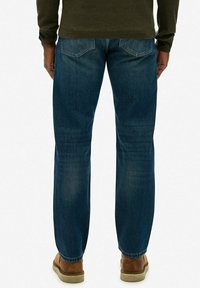 Niebieskie dżinsy o prostym kroju, z tylnymi kieszeniami z subtelnym detalem w kształcie litery V, noszone z brązowym obuwiem.