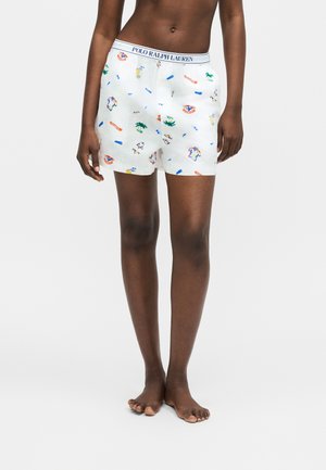 Mørkhudet person iført hvide Polo Ralph Lauren boxershorts med farverige små print, stående barfodet på en ensfarvet baggrund.