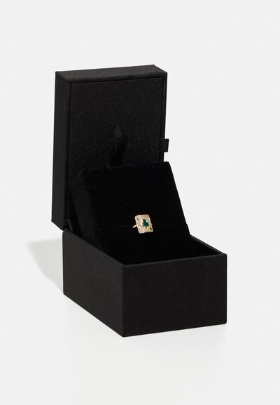 Caja de joyas negra parcialmente abierta, revelando un anillo dorado con un diseño rectangular, que presenta una piedra preciosa verde y pequeños acentos claros.