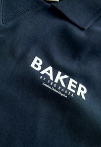 Marineblå polo shirt lavet af tekstureret stof med et hvidt logo "BAKER by TED BAKER" og lokationsdetaljer i mindre tekst nedenunder.