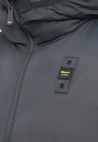 Giacca imbottita grigia con colletto alto, dotata di cerniera visibile e un'etichetta nera con testo giallo. Tessuto dalla texture liscia, design minimalista.