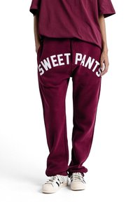 Pantalons de survêtement bordeaux avec "SWEET PANTS" imprimé en blanc. Coupe décontractée, texture douce, avec une taille à cordon. Associés à des baskets blanches.