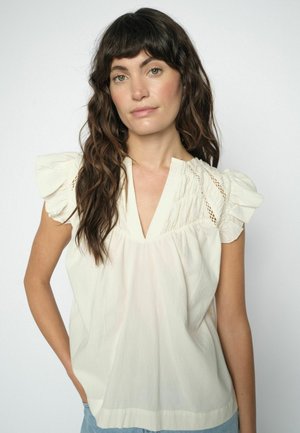 Žena s dlhými vlnitými hnedými vlasmi, oblečená v krémovej blúzke s volánikovými krátkymi rukávmi a výstrihom do V, stojaca pred jednoduchým svetlým pozadím.