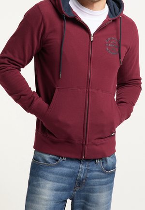 Mann trägt einen bordeauxroten Kapuzenpullover mit Reißverschluss und marineblauer Kapuzeninnenseite, bedrucktem Text auf der Brust, Hände in den Taschen, kombiniert mit blauen Jeans.