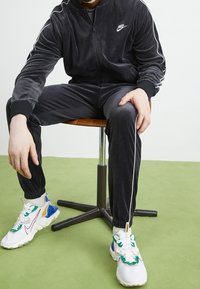 Svart velour-träningsdräkt med vita sidostråk, åtsittande ärmslut och jacka med dragkedja. Vita sneakers med färgglada detaljer och strukturerade sulor.