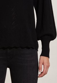 Pull en maille noir avec un motif en diamant texturé, des poignets côtelés et un ourlet festonné. Porté sur un jean noir, mettant en valeur une silhouette ajustée.