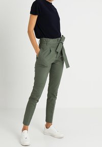 Gröna skräddarsydda byxor med hög midja, knytbälte och sidofickor, i kombination med en svart t-shirt med kort ärm och vita sneakers.