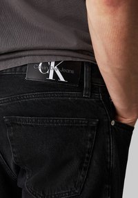 Čierne denimové džínsy s viditeľnou zadnou vreckom, so štítkom z čiernej kože s nápisom "Calvin Klein Jeans" a bielym logom.