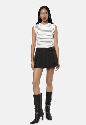 Kvinde stående iført hvid ærmeløs top med tynde sorte striber, sort plisseret mininederdel med bælte og sorte knæhøje støvler.