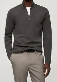 Man in een donkergrijze kwart-rits trui over een wit shirt, beige broek en zwarte riem, staand met één hand in zijn zak.