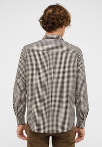 Mann mit lockigem braunem Haar, der ein kariertes Langarmhemd in Schwarz-Weiß und braune Hosen trägt, steht mit dem Rücken zur Kamera.